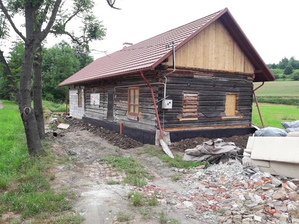 Stary drewniany dom z nową czerwoną dachówką i orynnowaniem, częściowo ocieplony, z widocznymi śladami prac ziemnych wokół fundamentów i stertą gruzu z przodu.