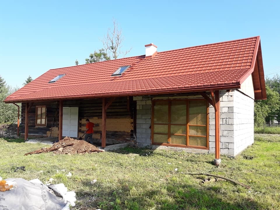 Dom w trakcie remontu z nowym, czerwonym dachem z dachówki, widoczne okna dachowe, fragmenty drewnianej konstrukcji i murowanej ściany, pracownik z łopatą.
