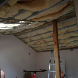 Montaż izolacji z wełny mineralnej w metalowym stelażu sufitu podwieszanego, widoczny fragment ściany i drabina.