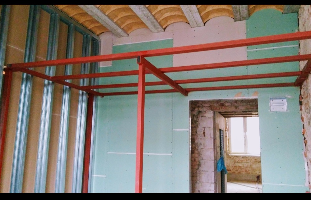 Wnętrze w trakcie budowy: czerwona stalowa konstrukcja sufitowa na tle zielonych płyt gipsowo-kartonowych, widoczne profile ścienne i fragment odsłoniętej cegły.