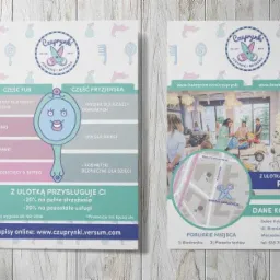 Dwie ulotki reklamowe salonu fryzjerskiego dla dzieci 'Czuprynki' leżące na drewnianym stole obok kubka z napojem i ołówka.