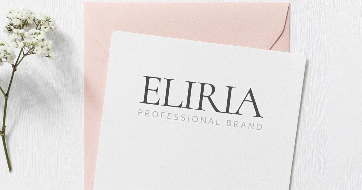 Karta z czarnym napisem 'ELIRIA PROFESSIONAL BRAND' leży obok jasnoróżowej koperty, całość dopełnia gałązka gipsówki na białym drewnianym tle.