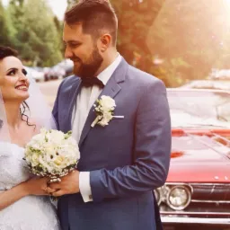 Uśmiechnięta para młoda w koronkowych strojach, trzymająca bukiet białych róż, pozuje na tle czerwonego, klasycznego samochodu w słoneczny dzień.