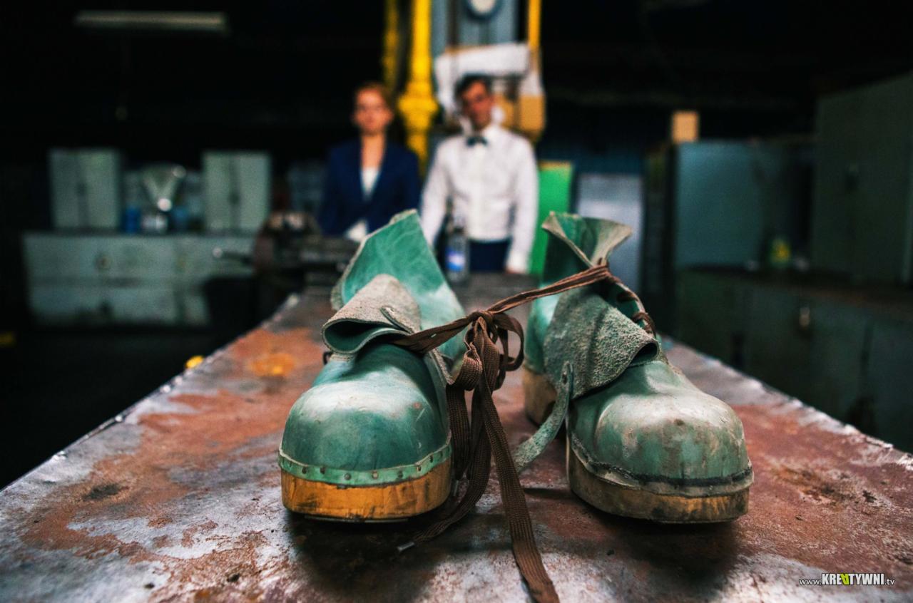 Dwa zielone, używane chodaki na drewnianej podeszwie, zawiązane brązowym sznurkiem, z parą w tle w warsztatowym otoczeniu.