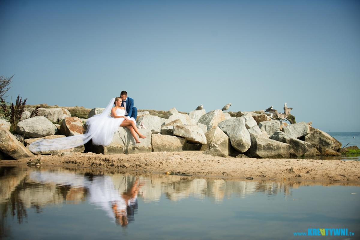 Para młoda siedząca na kamieniach nad brzegiem morza; panna młoda w białej sukni z długim welonem, pan młody w niebieskim garniturze, w tle mewy.