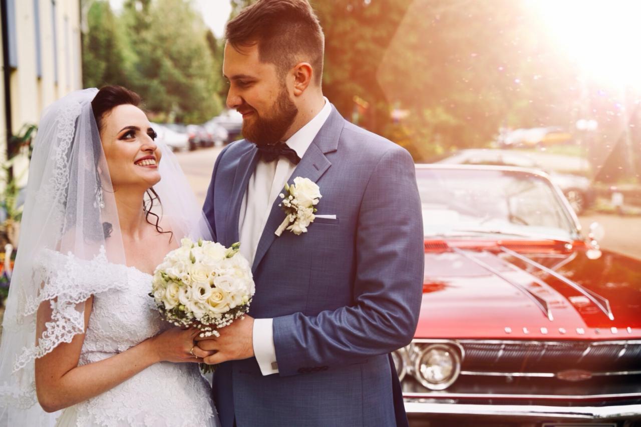 Uśmiechnięta para młoda w koronkowych strojach, trzymająca bukiet białych róż, pozuje na tle czerwonego, klasycznego samochodu w słoneczny dzień.