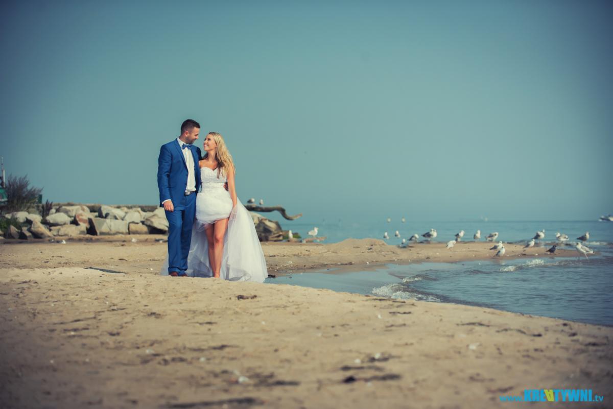 Para młoda na plaży, on w błękitnym garniturze z muszką, ona w krótkiej sukni ślubnej z długim trenem, w tle morze i mewy.