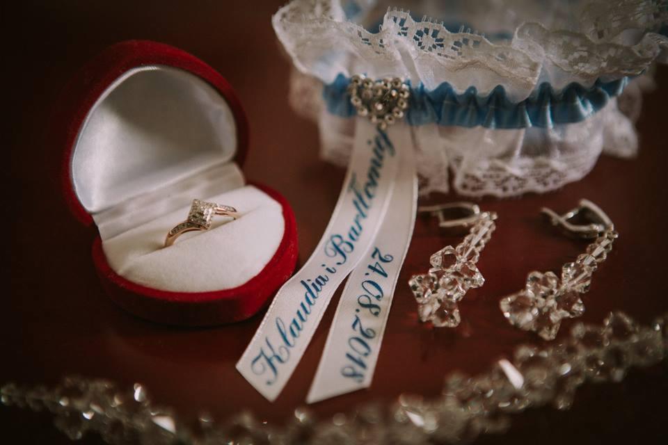 Złoty pierścionek zaręczynowy w czerwonym, aksamitnym pudełku, podwiązka ślubna z niebieską wstążką, kryształowe kolczyki i wstążka z imionami pary i datą ślubu na ciemnym tle.