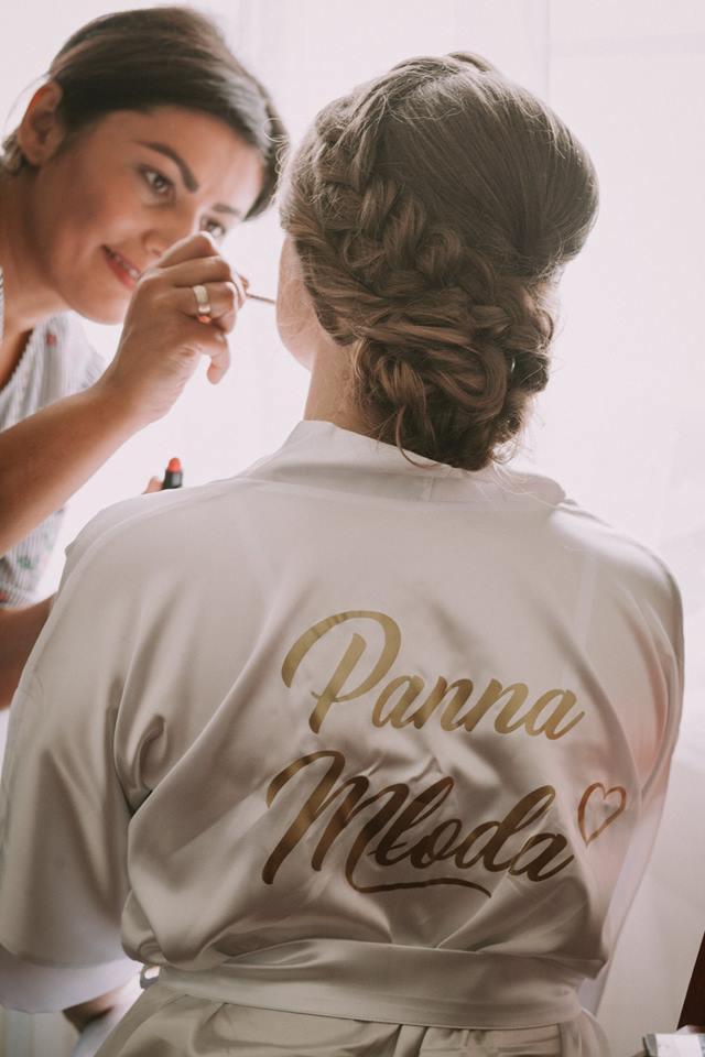 Wizażystka aplikuje makijaż pannie młodej, która ma na sobie satynowy szlafrok z napisem 'Panna Młoda' na plecach i upięte włosy