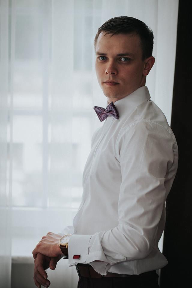 Portret mężczyzny w białej koszuli z bordową muchą i zegarkiem na tle okna z białymi zasłonami.