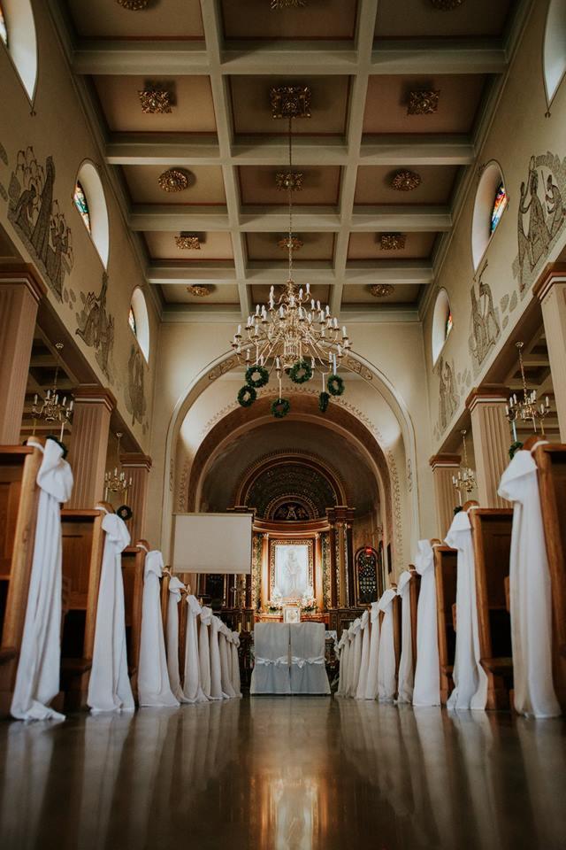 Uroczysta dekoracja kościoła na ślub: białe draperie na ławkach, kwiatowe girlandy na żyrandolu, ołtarz w tle.