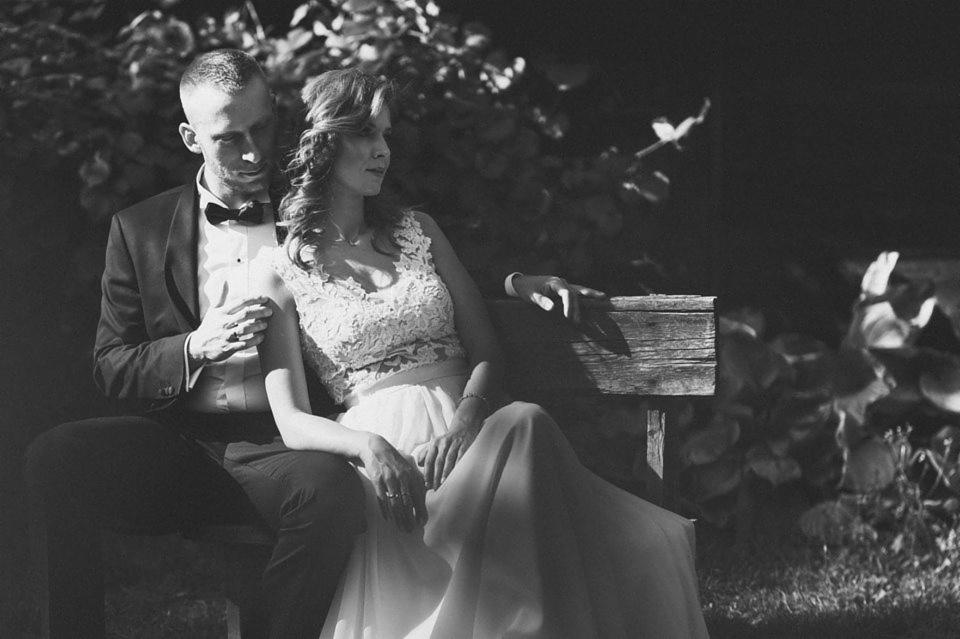 Czarno-białe zdjęcie pary młodej siedzącej na drewnianej ławce w parku, panna młoda w koronkowej sukience, pan młody w smokingu z muszką, delikatne światło słoneczne przebija się przez liście.