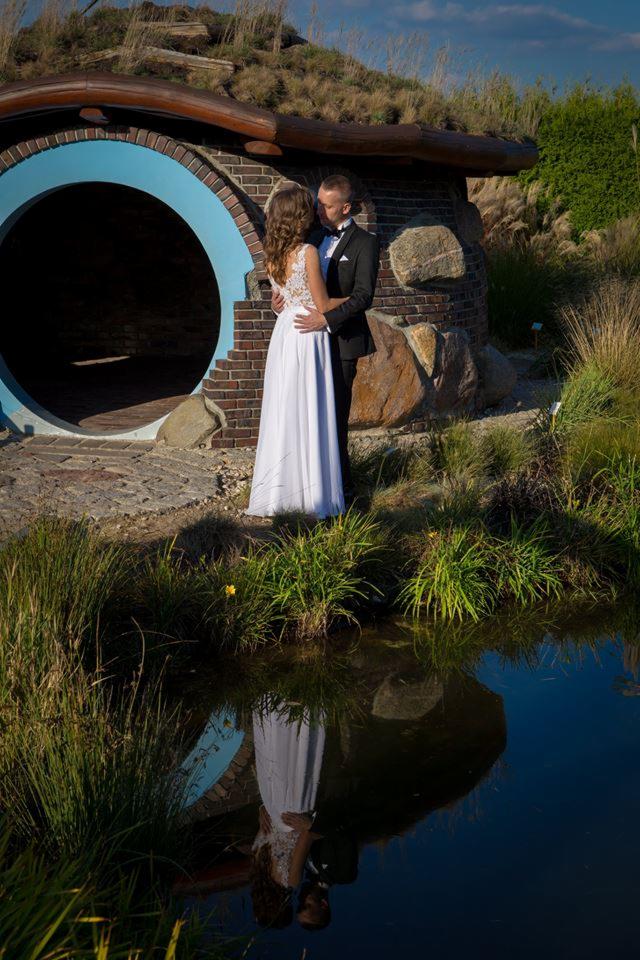 Para młoda w objęciach przed nietypową budowlą z cegły z okrągłym wejściem, odbicie w wodzie, sceneria w stylu Hobbiton.