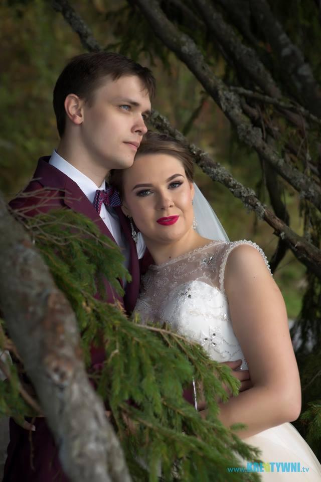 Para młoda w plenerze, panna młoda w białej sukni z koronkową górą i czerwonymi ustami, pan młody w bordowym garniturze i muszce, oboje stoją przy drzewie.