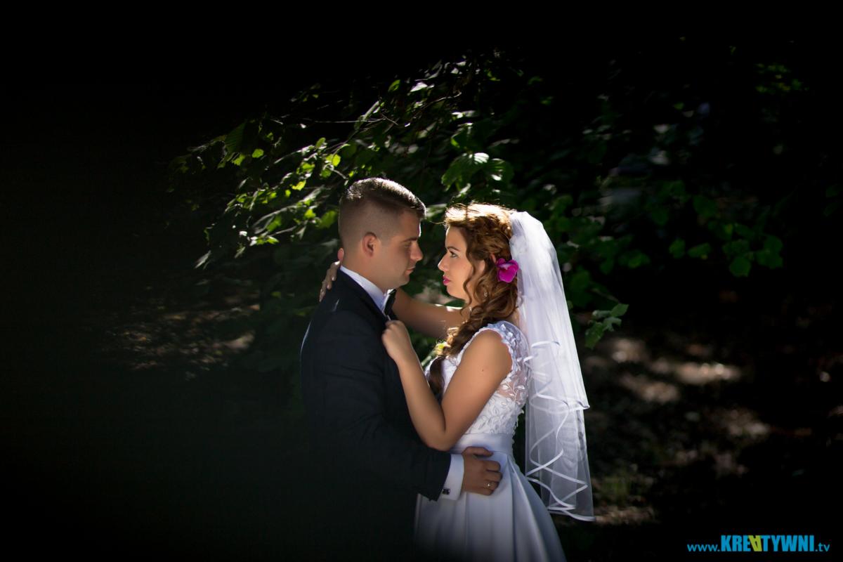 Para młoda w objęciach na tle ciemnej zieleni, panna młoda z różowym kwiatem we włosach, on w klasycznym czarnym garniturze.