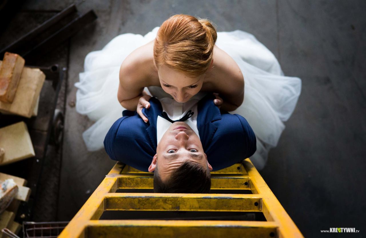 Nowożeńcy: panna młoda z rudymi włosami spogląda na pana młodego leżącego na żółtej drabinie, ujęcie z góry, industrialne tło.