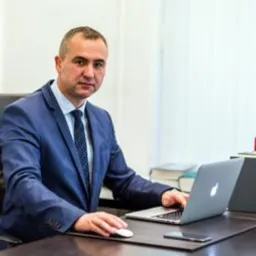 Mężczyzna w garniturze siedzi za biurkiem z laptopem, myszką i telefonem. W tle widoczne książki.