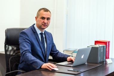 Mężczyzna w garniturze siedzi za biurkiem z laptopem, myszką i telefonem. W tle widoczne książki.