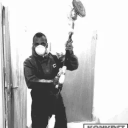 Pracownik w kombinezonie i masce szlifuje ścianę za pomocą szlifierki teleskopowej z odkurzaczem, widoczny fragment logo firmy 'KONKRET BUD' w prawym dolnym rogu.