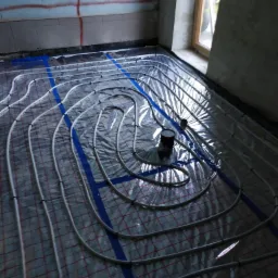 Instalacja ogrzewania podłogowego z rur w nowym budynku, widoczne nieotynkowane ściany i plastikowa folia ochronna z siatką.