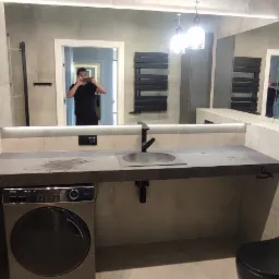 Nowoczesna łazienka w szarościach z pralką pod umywalką, widoczna kabina prysznicowa i duże lustro z odbiciem osoby robiącej zdjęcie.