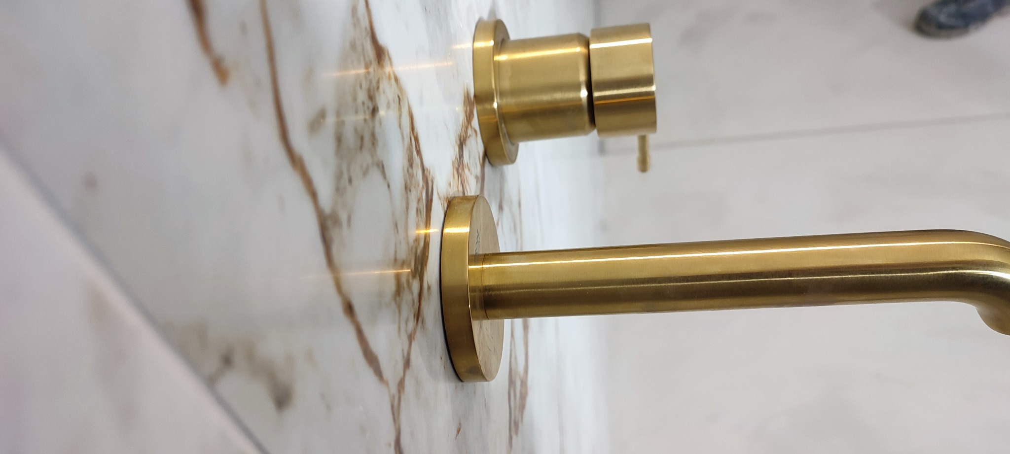 Bateria umywalkowa w kolorze złotym, zamontowana na ścianie z marmurowym wzorem, widoczny fragment podłogi i buta w tle.