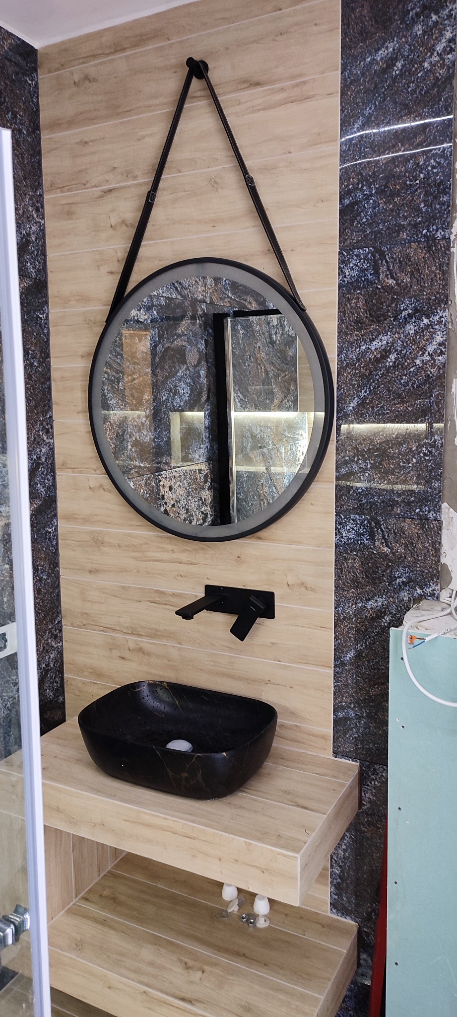 Częściowo wykończona łazienka z czarną umywalką nablatową, okrągłym lustrem na pasku i płytkami imitującymi drewno oraz kamień