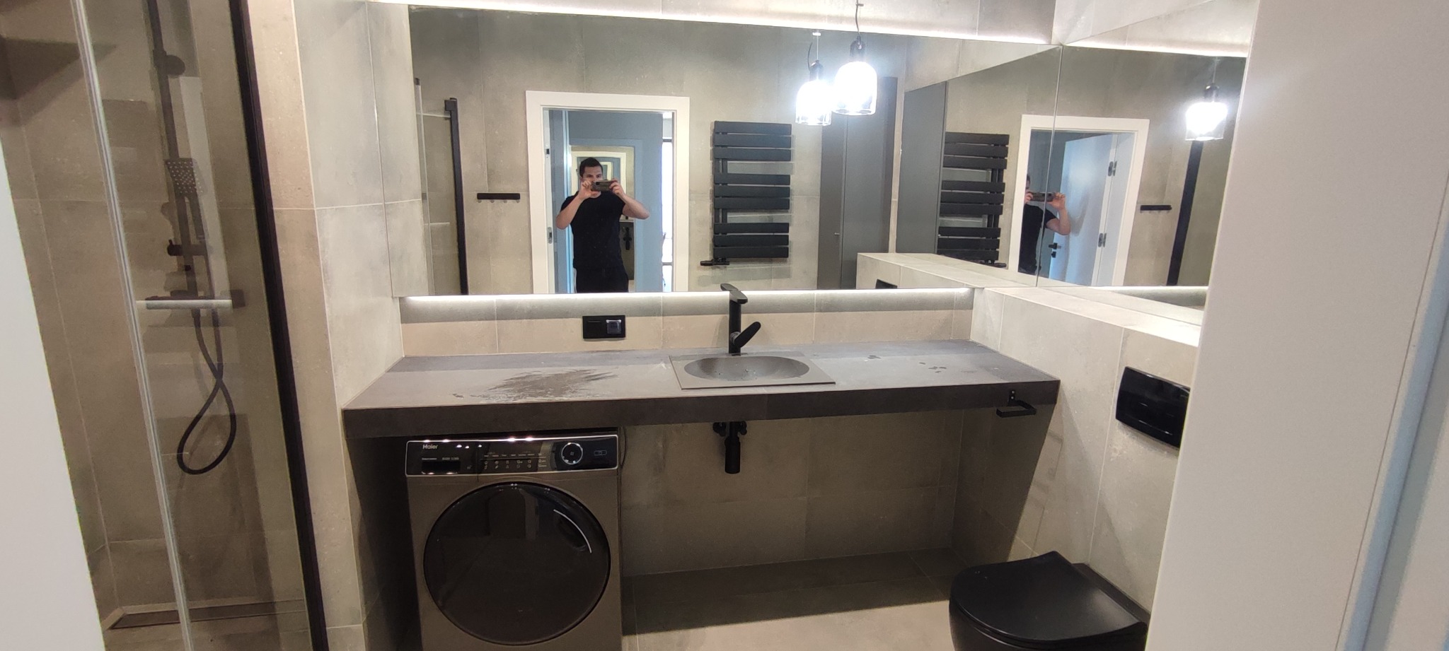 Nowoczesna łazienka w szarościach z pralką pod umywalką, widoczna kabina prysznicowa i duże lustro z odbiciem osoby robiącej zdjęcie.