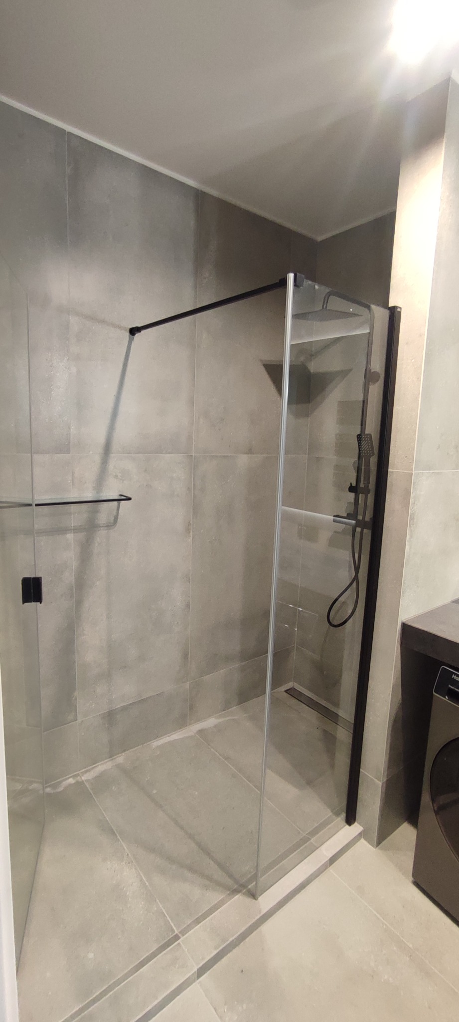 Kabina prysznicowa typu walk-in z czarnymi profilami i deszczownicą, zamontowana w łazience wykończonej szarą, dużą płytką imitującą beton.