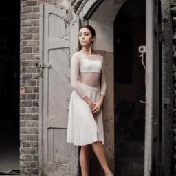 Młoda balerina w białym stroju tanecznym pozuje w otworze drzwiowym starej, ceglanej budowli, zamyślona twarz skierowana w lewo, delikatne światło podkreśla strukturę cegieł.