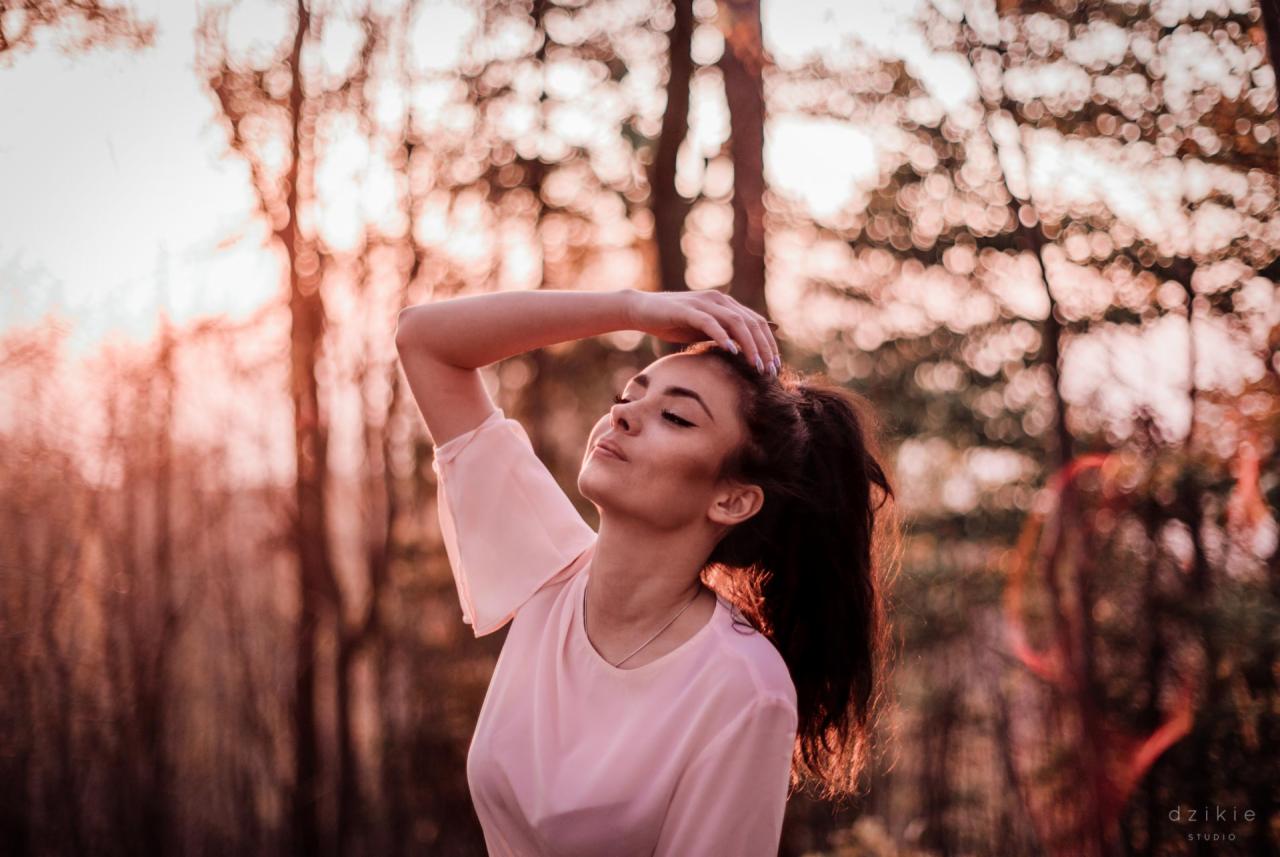 Kobieta w różowej bluzce, z zamkniętymi oczami i uniesioną ręką, pozuje na tle rozmytych drzew w ciepłym, zachodzącym słońcu.