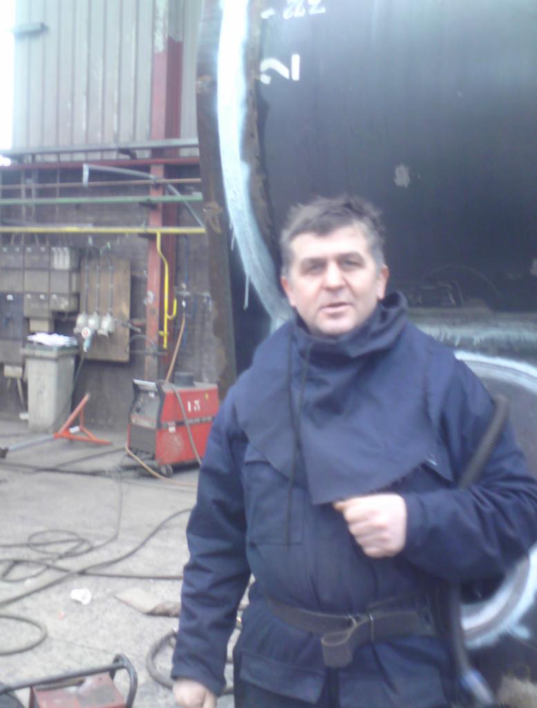 Mężczyzna w roboczym kombinezonie ochronnym stoi obok dużej, spawanej rury z widocznymi śladami spawania, na tle warsztatu z przewodami i urządzeniami spawalniczymi.