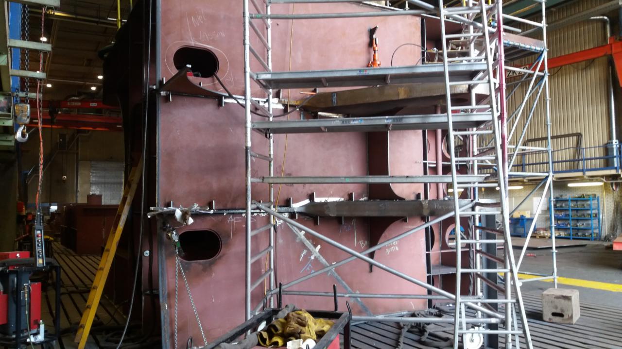 Konstrukcja stalowa z otworami, wsparta rusztowaniem, widoczne ślady spawania i montażu w hali produkcyjnej.
