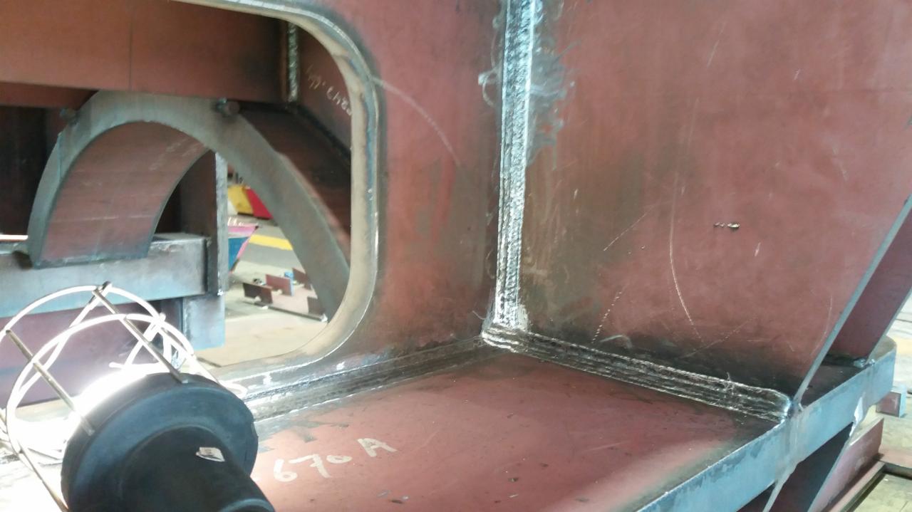 Czerwona, metalowa konstrukcja spawana z widocznymi śladami spawania i numerem '67-A' napisanym na powierzchni, oświetlona lampą warsztatową w klatce.