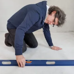 Mężczyzna w okularach, klęczący na posadzce, sprawdza poziom niebieską poziomicą w szarym pomieszczeniu z surowymi ścianami.