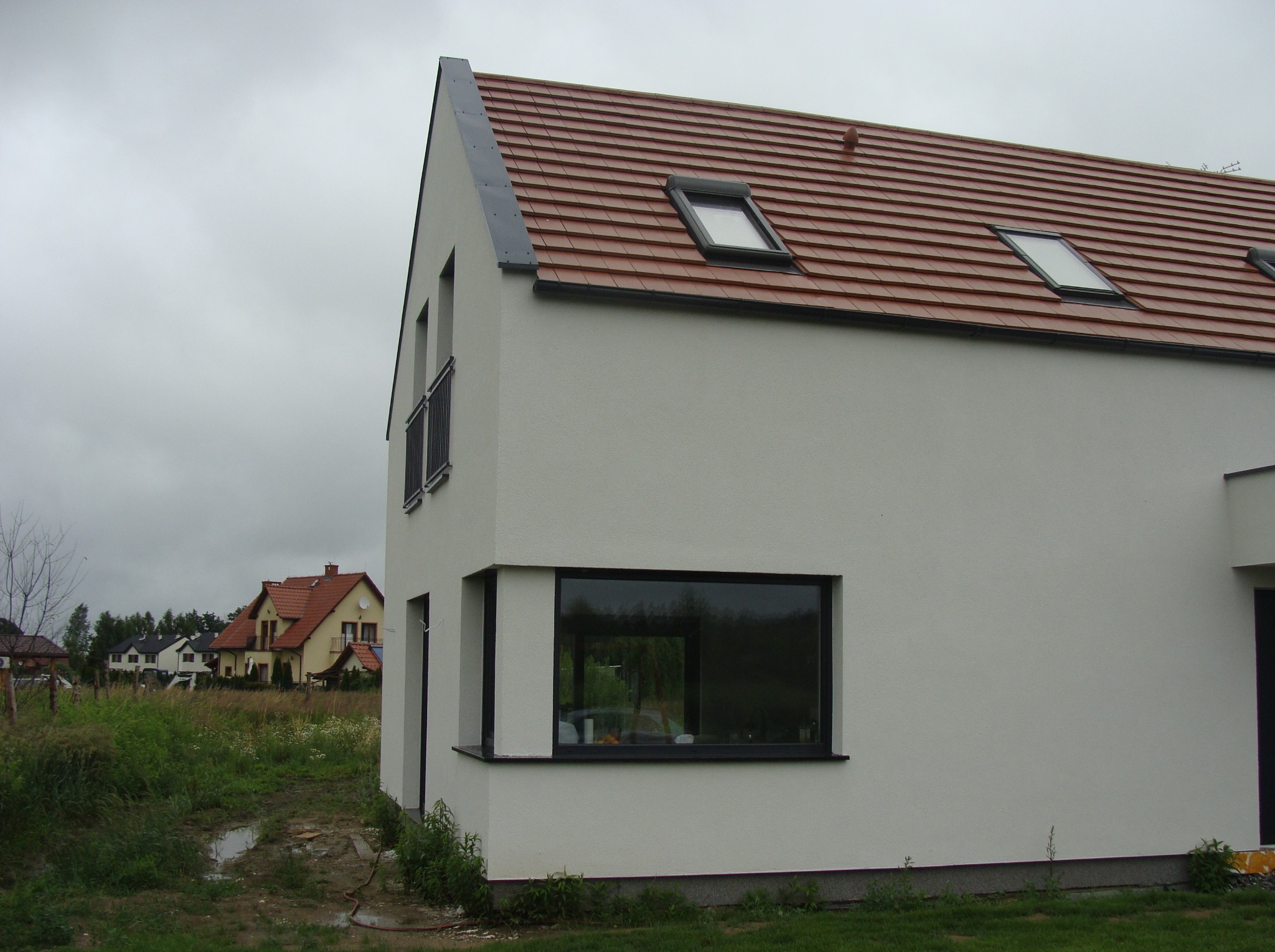 Nowoczesny, biały dom jednorodzinny z czerwoną dachówką i oknami dachowymi. Widok z zewnątrz, w tle inne domy i pochmurne niebo. Minimalistyczny design.