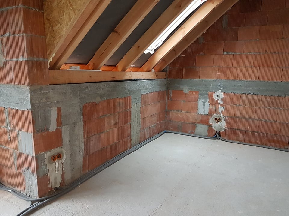 Surowe wnętrze poddasza z oknem dachowym, widoczne ceglane ściany, betonowy wieniec i instalacja elektryczna. Stan surowy zamknięty przed wykończeniem.