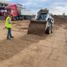 Niwelacja terenu: operator w kamizelce odblaskowej wyrównuje żwirowe podłoże minikoparką, w tle czerwona ciężarówka i hałda żwiru.