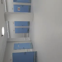 Korytarz szpitalny z rzędem niebieskich drzwi, białymi ścianami i podłogą, widoczne oświetlenie sufitowe