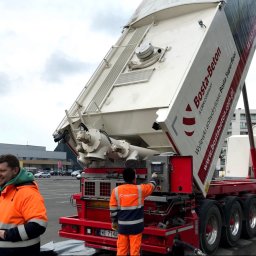 VAK SPÓŁKA Z OGRANICZONĄ ODPOWIEDZIALNOŚCIĄ - Specjalistyczna ciężarówka Bosta-Beton z podniesionym zbiornikiem do wylewek anhydrytowych, obsługiwana przez pracownika w pomarańczowej kurtce, na tle parkingu i budynków.