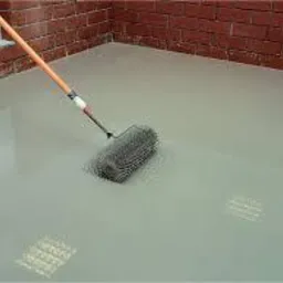 Aplikacja wylewki anhydrytowej wałkiem kolczastym na gładkiej, szarej powierzchni posadzki, widoczna ceglana ściana w tle.