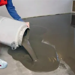 Wylewanie szarej, płynnej masy z plastikowego pojemnika na podłogę w pomieszczeniu z grzejnikiem i oknem w tle; widoczne fragmenty czerwonych spodni i sportowych butów osoby wylewającej.