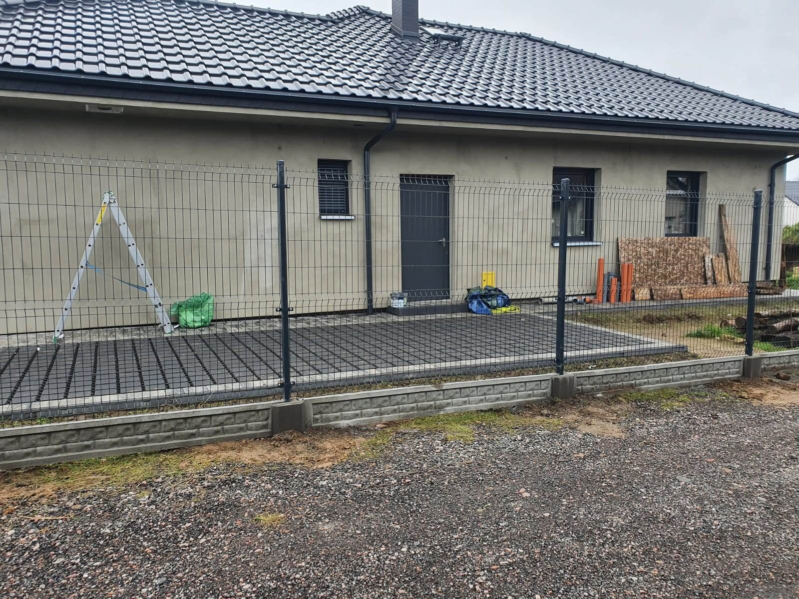 Nowoczesne, grafitowe ogrodzenie panelowe z podmurówką na tle domu jednorodzinnego, widoczne narzędzia pracy i materiały budowlane.