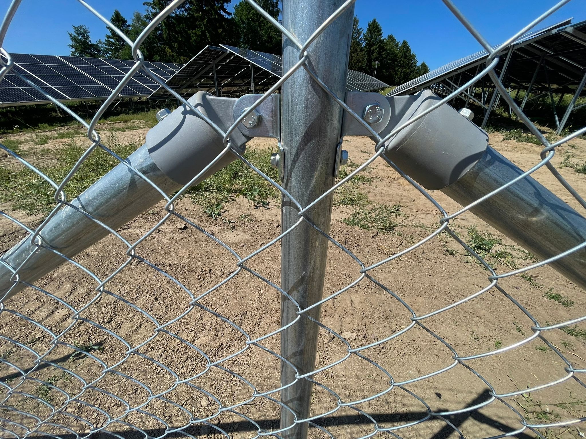 Metalowy słupek z mocowaniem i siatką ogrodzeniową na tle paneli słonecznych i drzew
