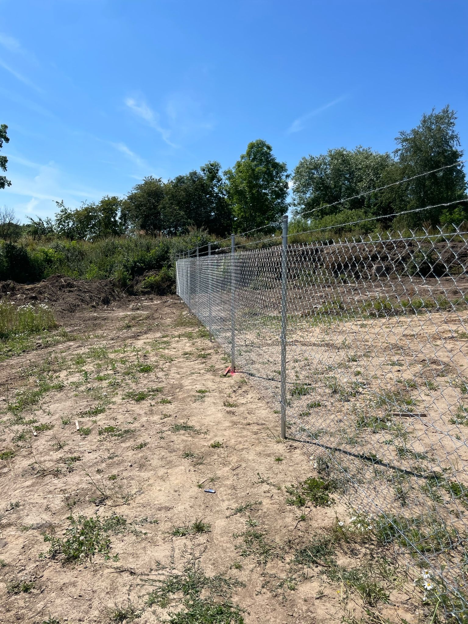 Długa linia srebrnej, metalowej siatki ogrodzeniowej z drutem kolczastym u góry, rozciągająca się przez piaszczysty teren z kępami zieleni i drzewami w tle, pod błękitnym niebem.