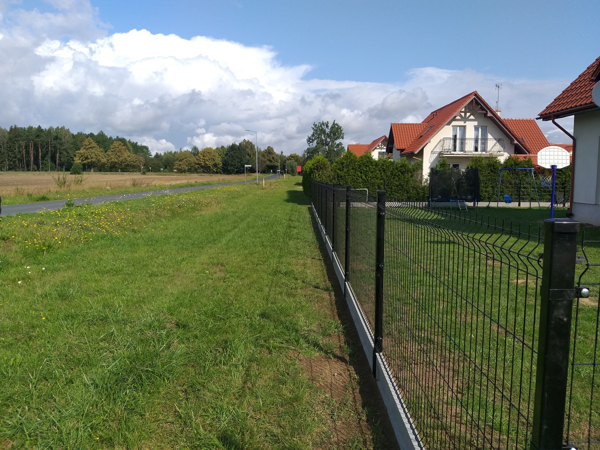 Długi, czarny panel ogrodzeniowy z metalowej siatki, rozciągający się wzdłuż zielonego trawnika, z domami w tle pod błękitnym niebem z chmurami.