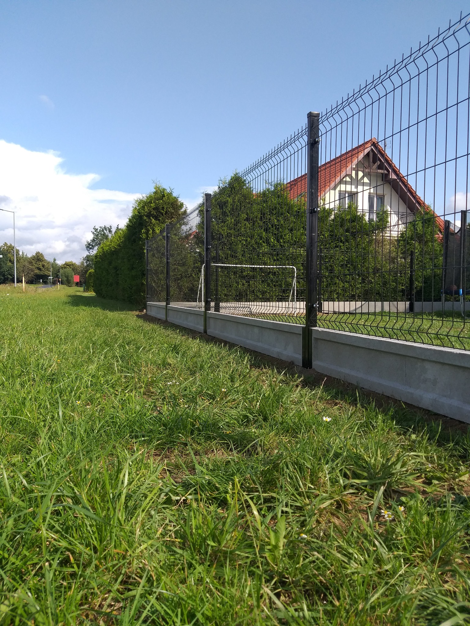 Nowoczesne, czarne ogrodzenie panelowe z podmurówką betonową, widok z poziomu trawy, w tle dom z czerwonym dachem i zielone tuje.