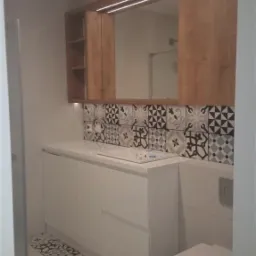 Nowoczesna łazienka z białą szafką pod umywalką, drewnianymi szafkami wiszącymi z oświetleniem LED, dekoracyjnymi płytkami na ścianie i podłodze.