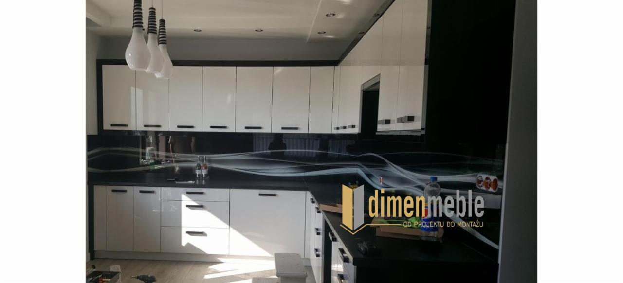 Biała zabudowa kuchenna z czarnym blatem i dekoracyjnym panelem szklanym nad blatem, oświetlona wiszącymi lampami. Widoczne narzędzia i logo firmy na blacie.