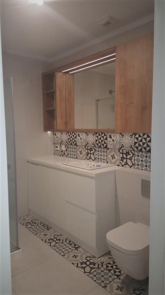 Nowoczesna łazienka z białą szafką pod umywalką, drewnianymi szafkami wiszącymi z oświetleniem LED, dekoracyjnymi płytkami na ścianie i podłodze.